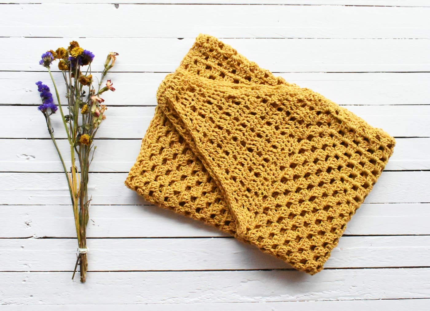 A folded mustard gold crochet blanket on a white wood table with dried flowers